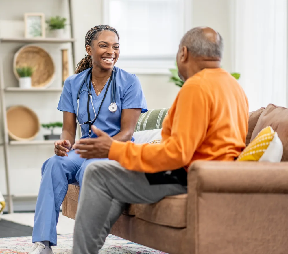 A care coordinator in scrubs laughing with an older patient on his sofa