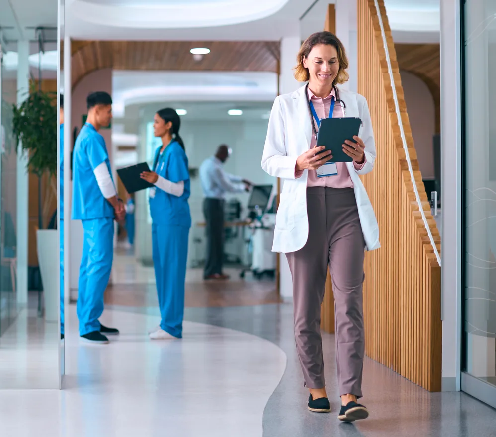 A doctor with a tablet walking down a hospital corridor past two nurses