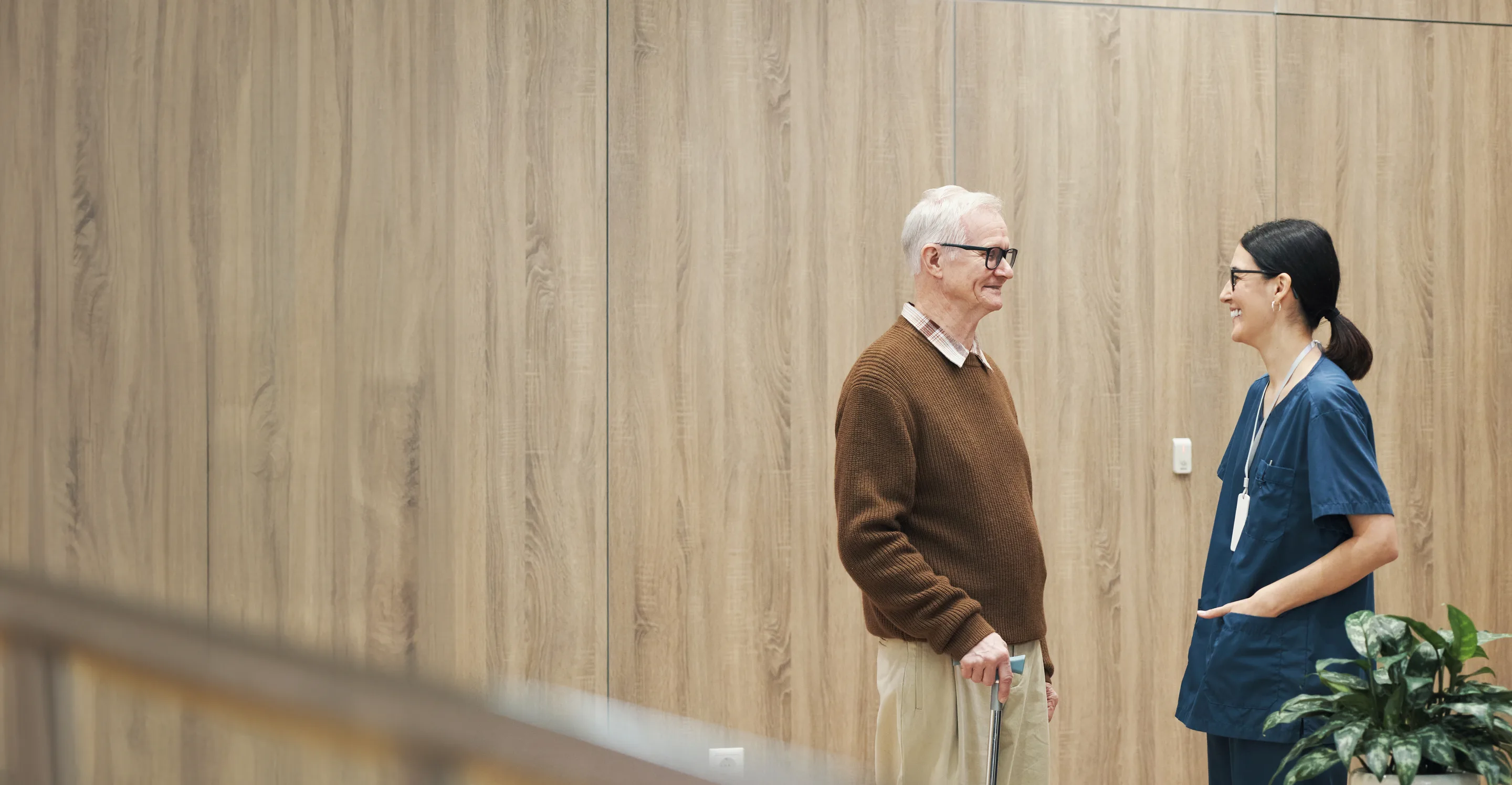 An older patient with a cane talking with a smiling nurse in a clinic hallway
