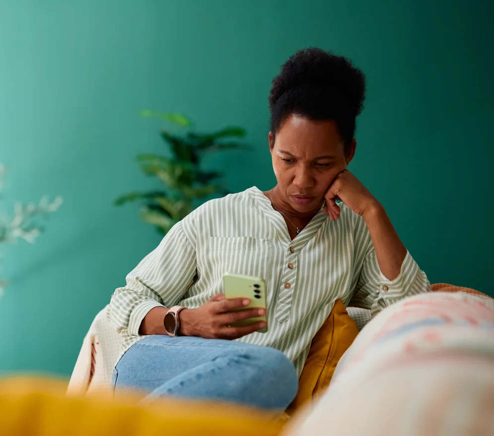 A woman sitting at home on a sofa, reading her phone with a worried expression