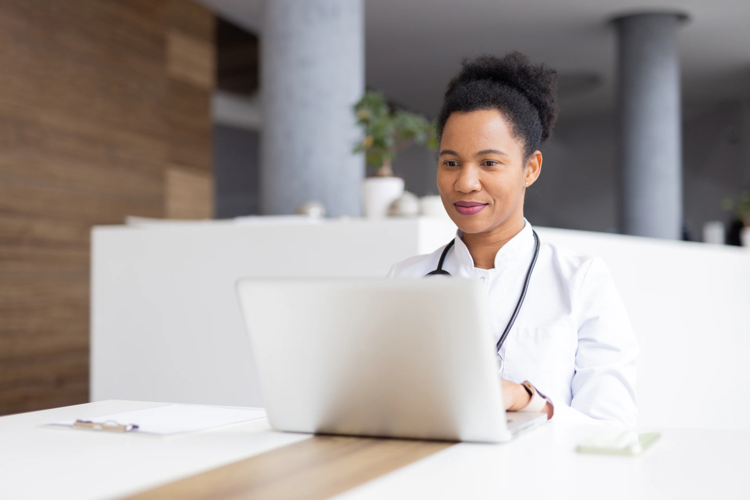 A clinician in a white coat working at a laptop in a bright clinic