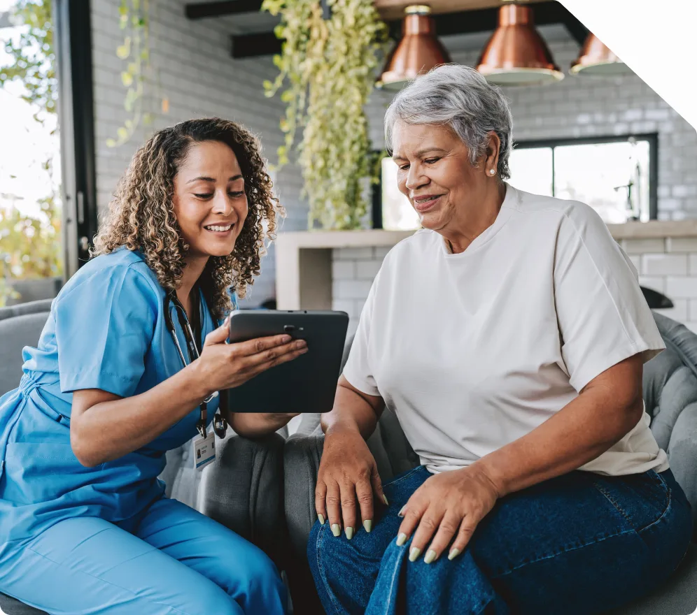 A nurse in scrubs showing a tablet to a seated patient at home