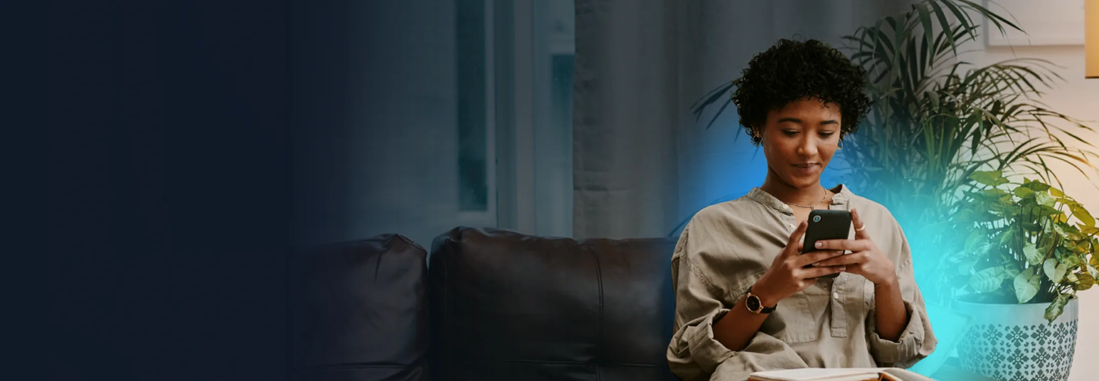 A woman sitting on a sofa checking her phone, lit by a glowing houseplant beside her