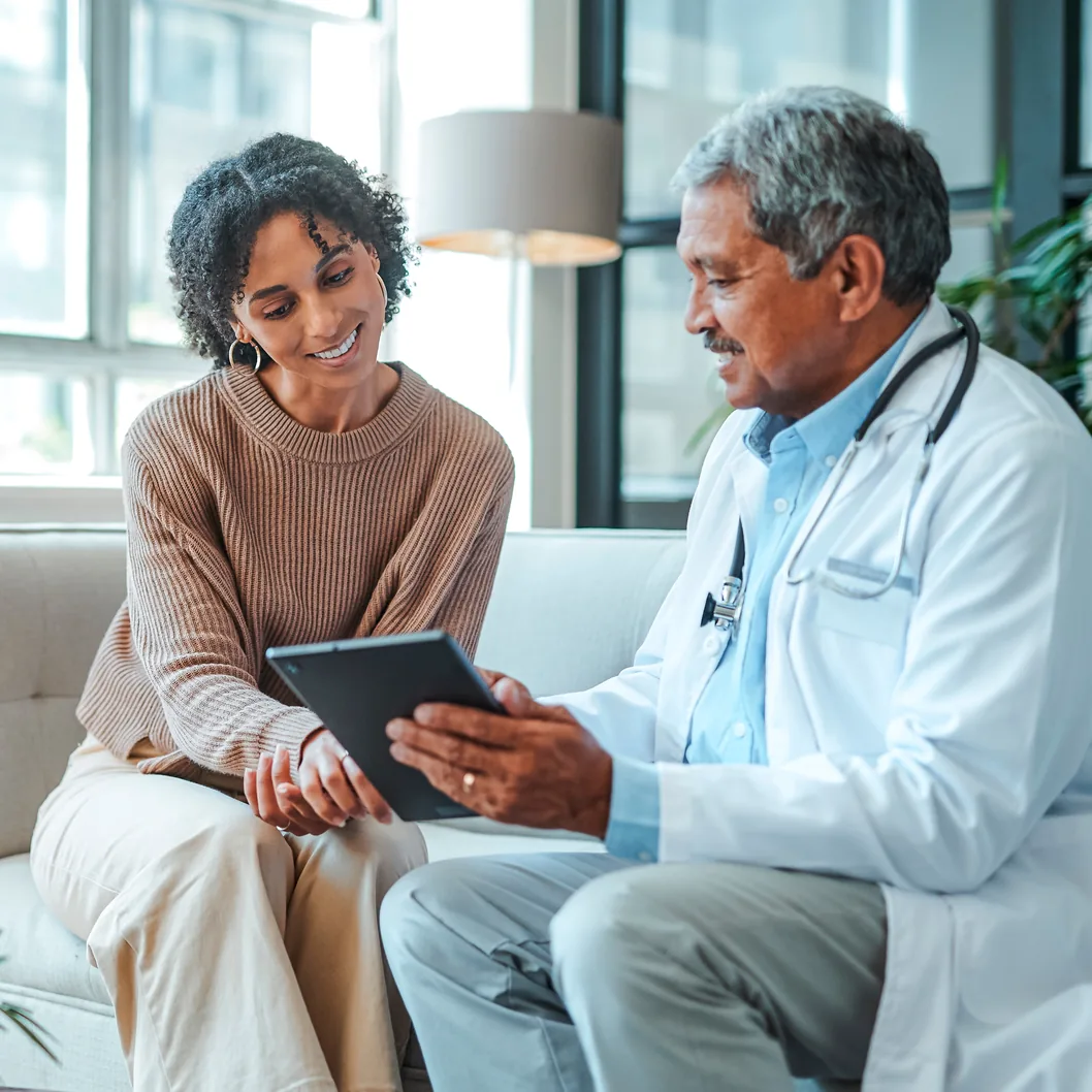 A patient and a clinician in a white coat sitting on a sofa, looking at a tablet together