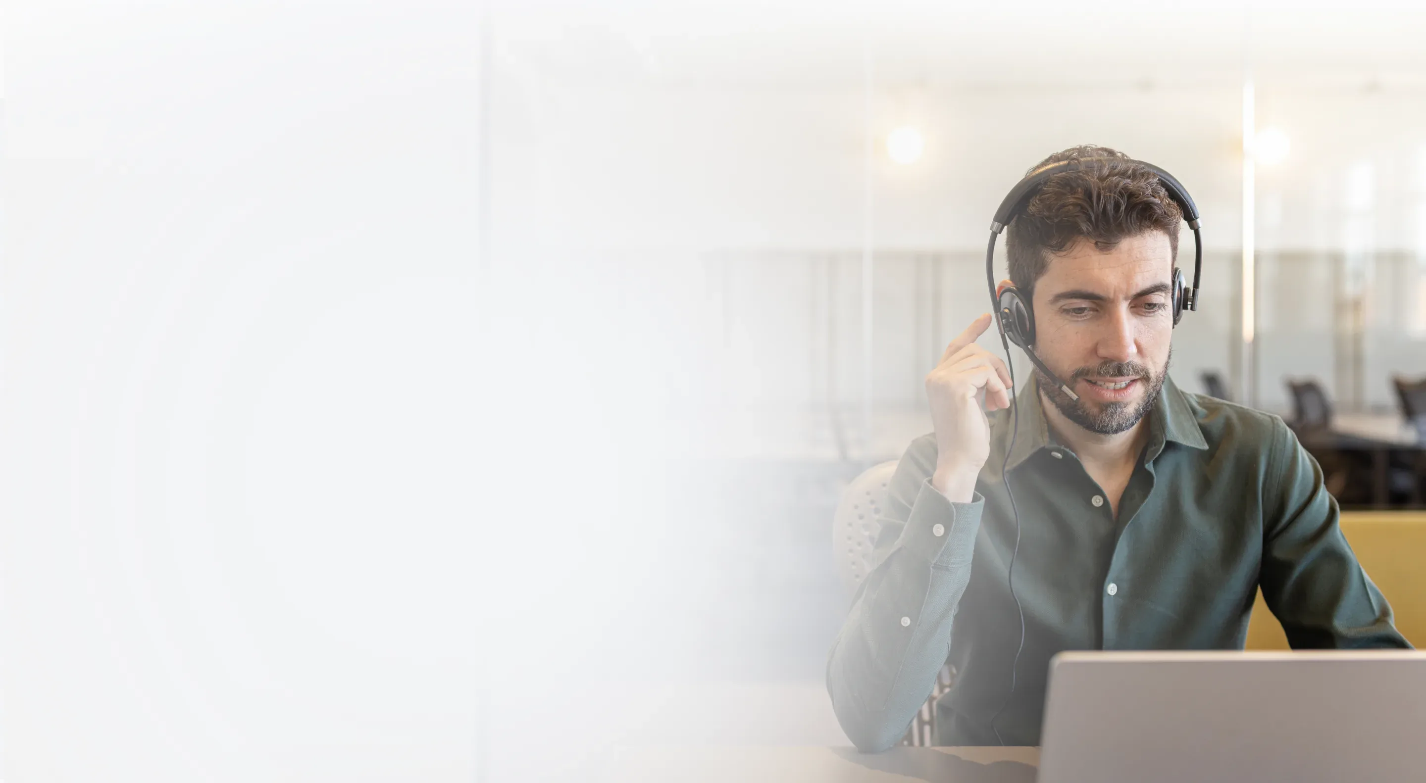 A man wearing a phone headset working at a laptop in an open-plan office