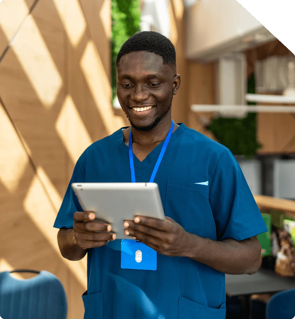 A nurse in blue scrubs smiling at a tablet in a sunlit clinic