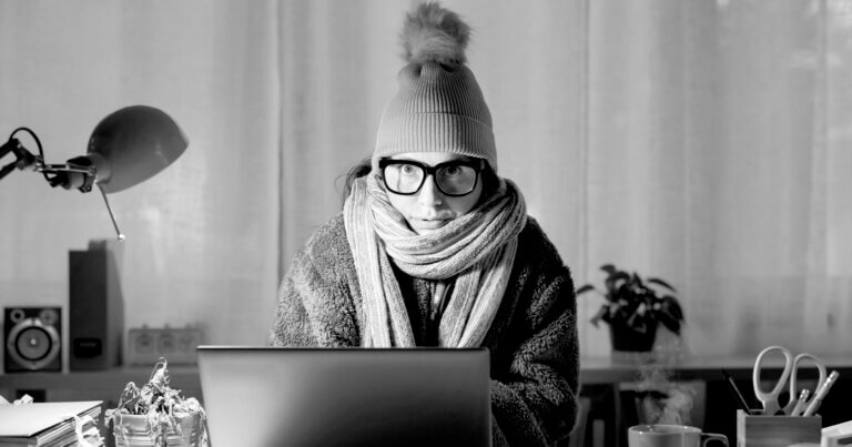 Woman bundled in winter weather gear sits at a laptop, looking overwhelmed