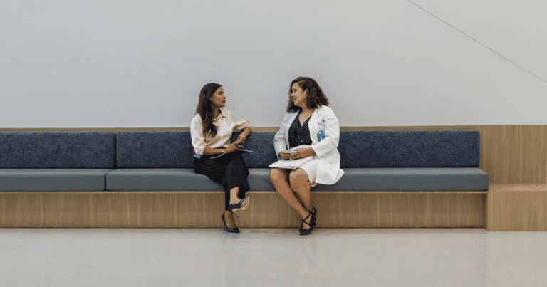 A photograph of a doctor and a patient sitting on a bench in a hospital setting.
